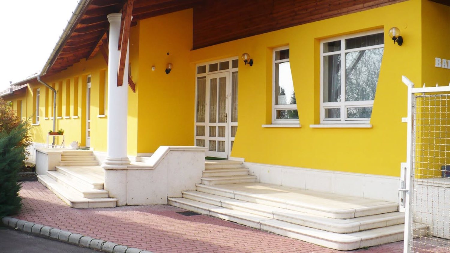 A hotel fedett bejárati előtere: fehér oszlop, faborítású eresz, világos kőlépcsők és üvegezett bejárati ajtó a sárga falban.